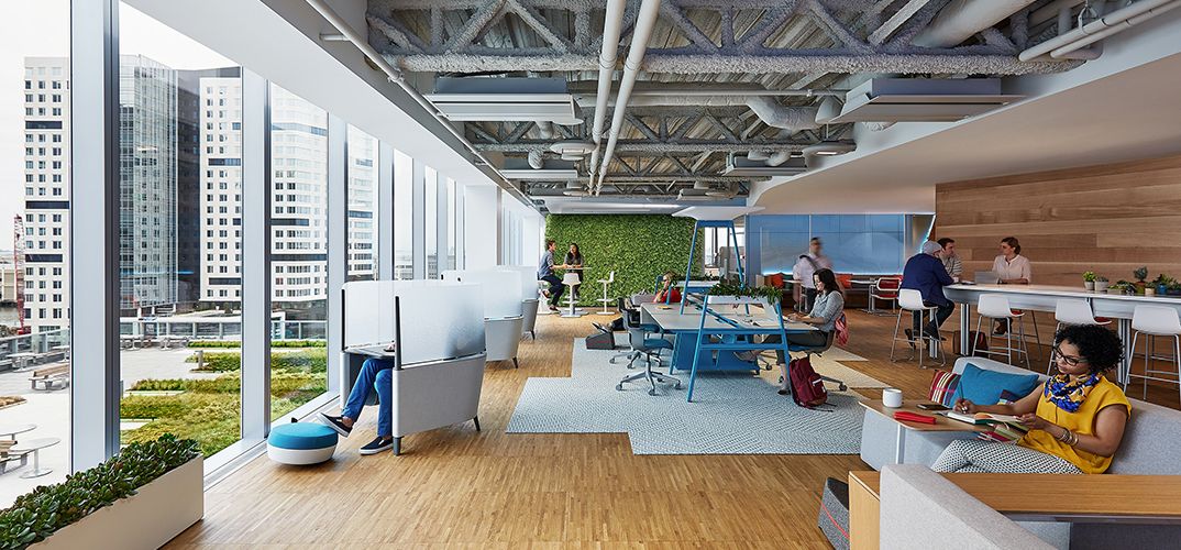 Light-filled, people-filled open workspace with privacy pods along the left window, collaboration space in the middle of the room and along the right wall.